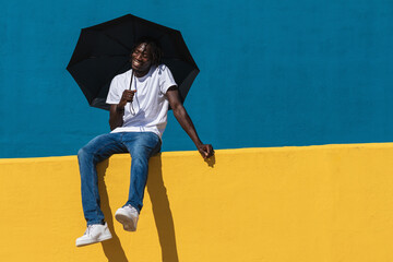 Happy black man holding an umbrella while sitting on yellow wall