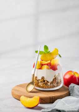 Greek yogurt parfait with peach and muesli in a glass on a wooden board on a light background with fresh fruits, napkin and shadow.
