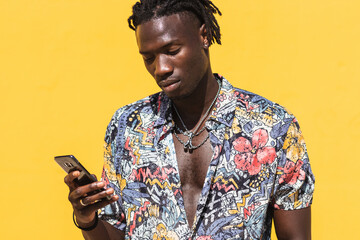 Portrait of African American man in colourful shirt isolated on yellow wall using mobile phone