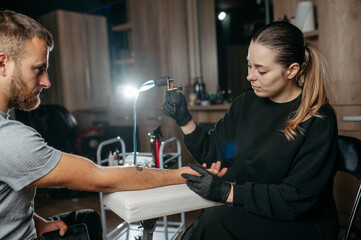 Young woman professional tattoo artist in black gloves making a rose tattoo on a man's forearm.