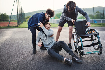 Young disabled man fall off wheelchair, fiends helping to sit back. Male friendship.