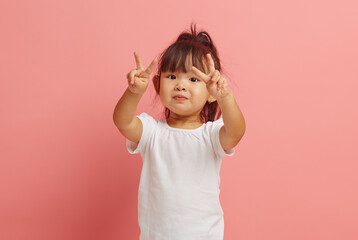 little korean etnicity girl showing V gesture with two hands standing over pink isolated...