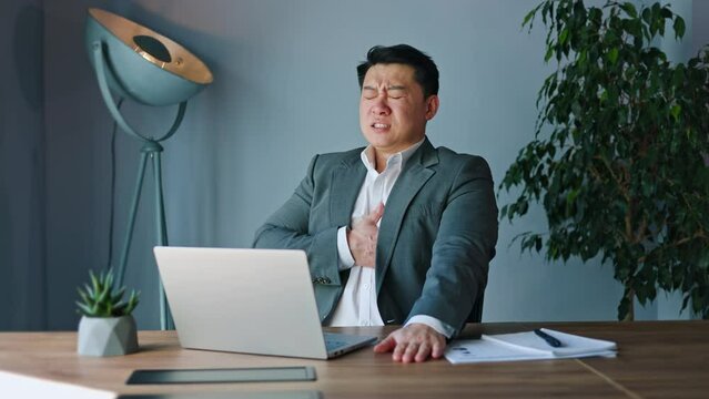 Troubled asian man clasping chest with pained expression and experiencing discomfort in heart. Sad young male doing work despite inner turmoil during demanding workday in sleek modern office.