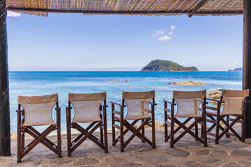 A row of chairs on the beach overlooking the Ionian sea with Turtle Island in the background,...