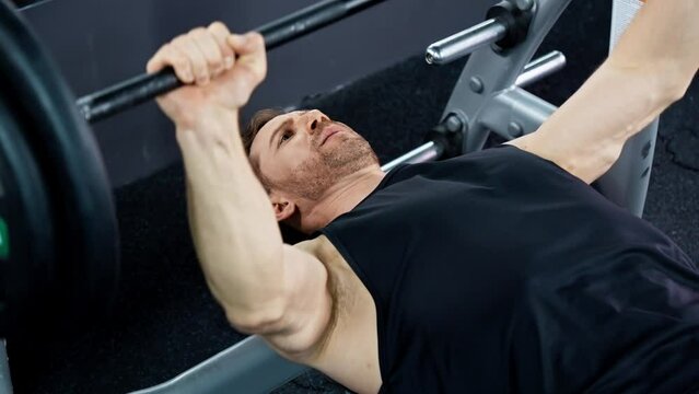 Athlete doing bench press workout with barbell in gym. Man lifting weights indoors. Chest training. Man is focused and determined. Building muscles indoors. Athlete in tank top. 4K, UHD