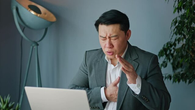 Unhealthy korean man having heart attack during workday while sitting at personal workplace in front of laptop in modern cabinet with decorative items and blue walls. Concept of pain and cardio.