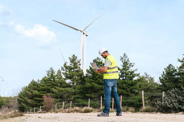 A technician in a hard hat and reflective vest works on a laptop near wind turbines on a dirt road.