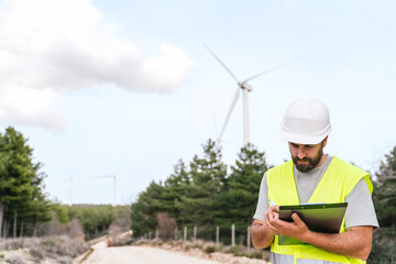 Fototapeta premium A wind farm technician checks notes on a clipboard, wearing a hard hat and safety vest, with wind turbines around.