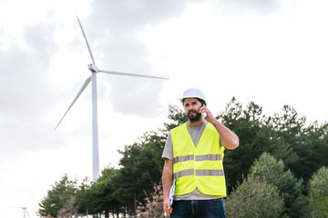 A wind turbine technician speaks on the phone, holding a clipboard, wearing a hard hat and safety vest in a wind farm.