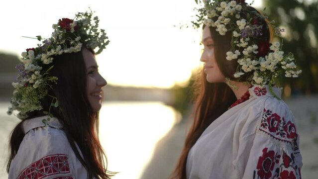 Slow motion. Rear view. Two Ukrainian women turn around and look at the camera, smiling. Women in traditional embroidered shirts with wreaths of wild herbs and flowers on their heads, stand the summer