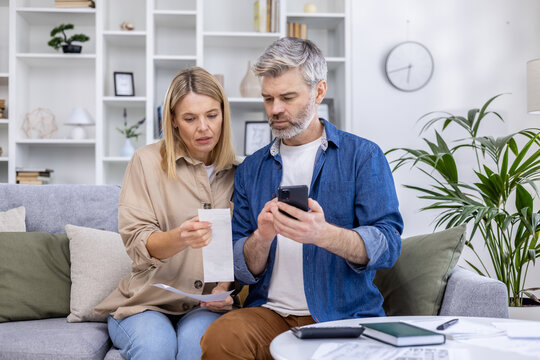 Middle-aged couple managing finances and reviewing household bills together at home while discussing their budget and using a smartphone. - Powered by Adobe
