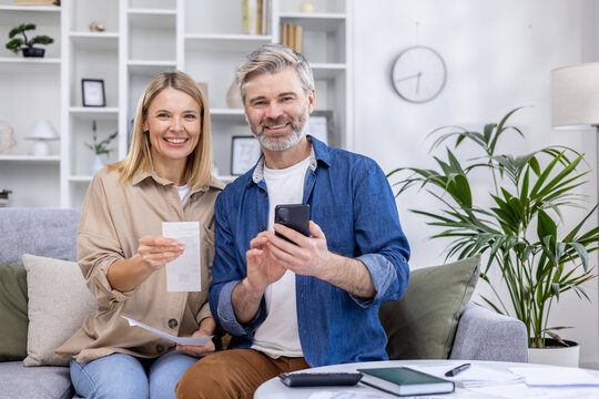 Happy mature couple using a smartphone for online banking while sitting on a sofa at home. They are handling finances together in a cozy living room. - Powered by Adobe