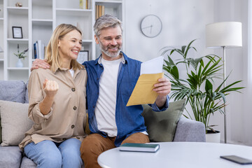 Happy couple celebrating with documents in hand at home. They are smiling and expressing excitement...