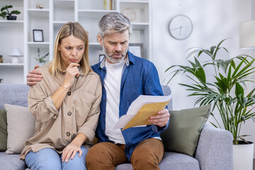 A concerned couple sitting on the couch, examining financial paperwork in their living room. They appear stressed while trying to understand the contents of the envelope.