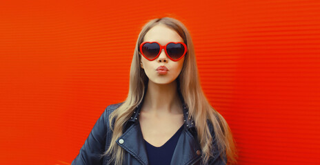 Portrait of beautiful stylish blonde young woman model blowing kiss in red heart shaped sunglasses