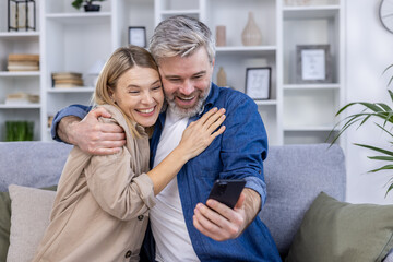 Joyful middle-aged couple hugging and taking a selfie on a smartphone while sitting on the couch in a cozy living room setting.