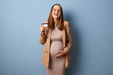 Laughing positive pregnant woman wearing dress and jacket looking at camera while stroking her...