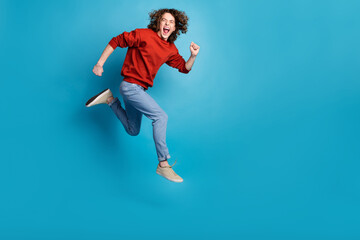 Full length photo of funny excited guy wear red sweater jumping high running emtpy space isolated blue color background