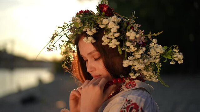 Slow motion. The girl adjusts the red ethnic necklace on her neck and looks thoughtfully. A young woman wearing a traditional embroidered shirt and a wreath of wild herbs and flowers on her head