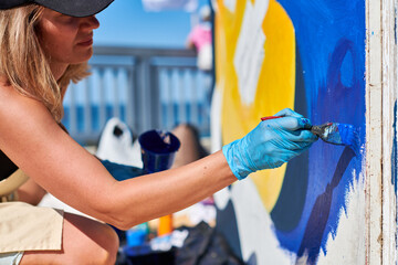 Adult female painter in black cap passionately paints picture with paintbrush for outdoor street...