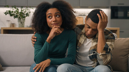 Annoyed African American ethnic exhausted young couple disturbed parents sitting sofa thinking...