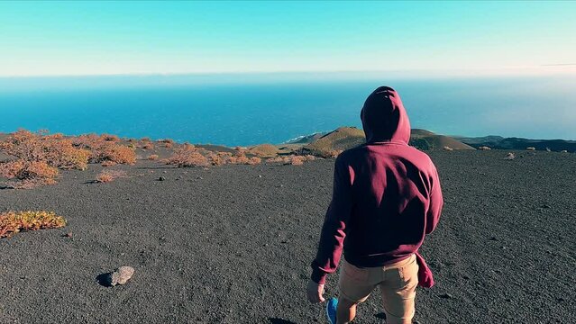 Person dressed in a red hooded sweatshirt walking on a volcanic area with the sea in the background on a sunny and windy day.