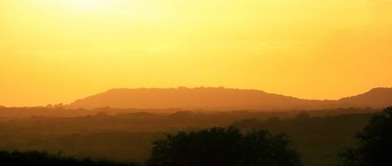 Orange sky with hues of pink, illuminating a mountain range with scattered trees and rolling hills