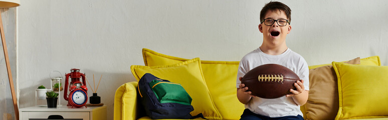 Little boy happily holds a football while sitting on a couch at home.