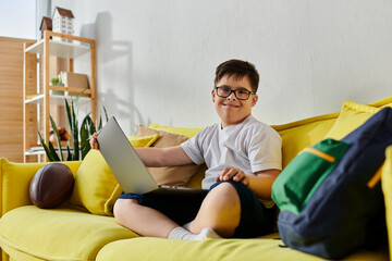 A adorable boy with Down syndrome sitting on a yellow couch, using a laptop.