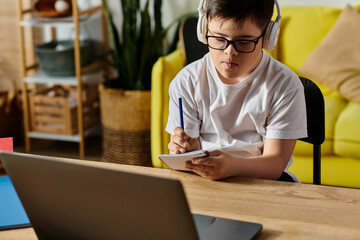 A adorable boy with Down syndrome is engrossed in using a laptop and wearing headphones at a table.