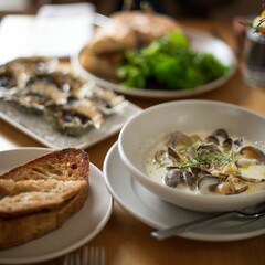 a bowl of creamy clam chowder, a freshly-baked piece of bread, oysters, and a sandwich on the table