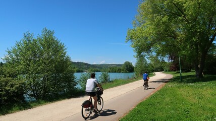 Cyclotourisme au bord du lac du chanoine Kir à Dijon, en Côte d’Or / Bourgogne, paysage de nature avec deux cyclistes roulant sur une piste cyclable au bord de l'eau (France) - obrazy, fototapety, plakaty