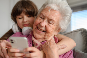 Grandmother with cute girl scrolling on smartphone, girl teaching senior woman to work with technology, internet. Portrait of elderly woman spending time with granddaughter.