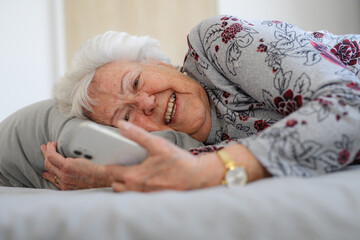 Senior woman lying in bed, looking at smartphone and smiling. Older woman using technology, digital skill and literacy.