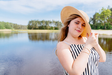 A woman wearing a straw hat and striped dress is standing beside a lake