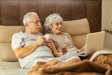 Loving senior couple sitting on a bed in a bedroom, enjoying coffee and using laptop for entertainment