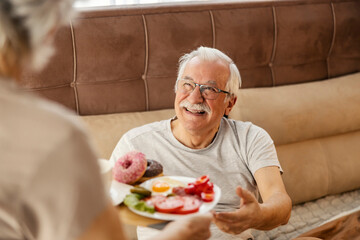 Smiling senior man is getting breakfast in bed at cozy bedroom in the morning.