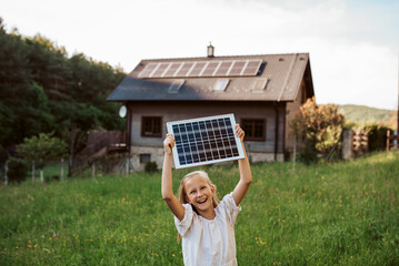 Little girl with model of solar panel, standing in the middle of meadow, house with solar panels behind. Concept of renewable resources.