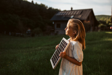 Little girl with model of solar panel, standing in the middle of meadow, house with solar panels behind. Concept of renewable resources.
