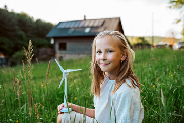 Girl with model of wind turbine, standing in the middle of meadow, house with solar panels behind. Concept of renewable resources.