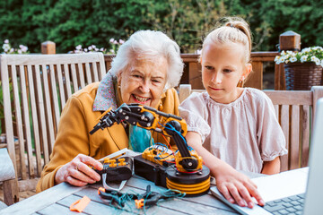 Grandmother supporting granddaughter in robotics, science, IT technologies and programming. Young girl assembling a simple robot and shoving it to grandma.