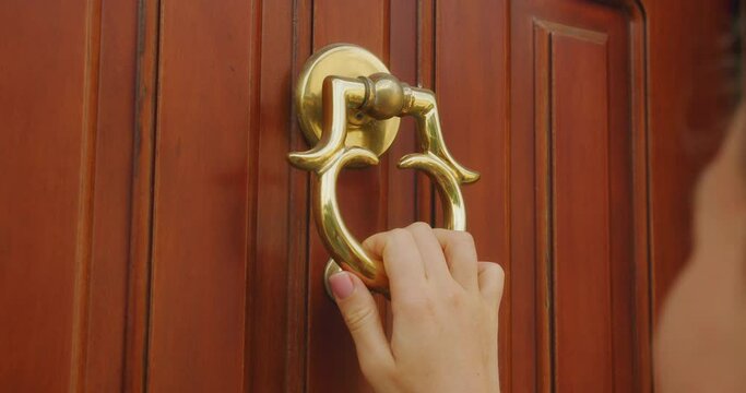 Hand holds ornate brass door knocker on a wooden door and knocks for the people living at home to open and let the wanderer in.