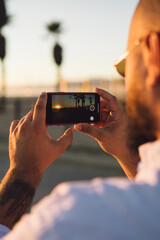 Close up back view of male holding mobile phone for shooting beautiful landscape of sundown during...