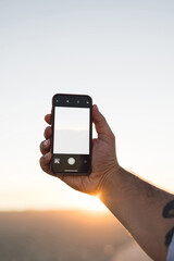 Cropped image of male using smartphone with blank copy space screen on background of beautiful sunset in evening, man's hand holding mobile phone with mock up area for web content or app on display