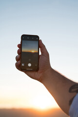 Cropped image of man's hand holding smartphone shooting video of evening sky and sundown, male using modern mobile phone camera taking photo of golden sunset  and nature during summer evening