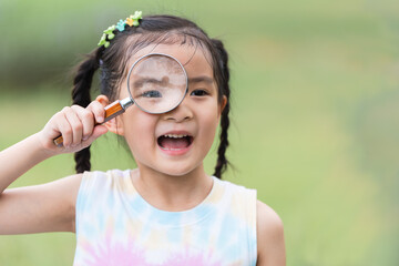 Portrait of Asian child girl in park playing outdoor in nature. Little girl standing, holding and looking through magnifying glass, smiling with curious face in casual clothes. Education
