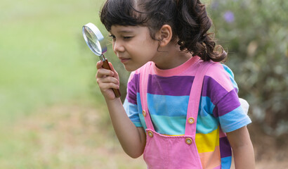 Portrait of Caucasian child girl in park playing outdoor in nature. Little girl standing, holding and looking through magnifying glass, curious smiling in casual clothes. Education. Side view