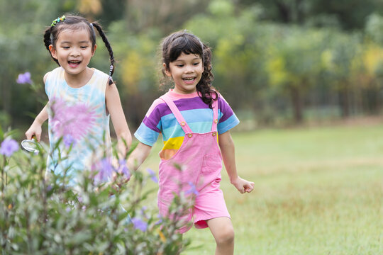 Asian and Caucasian little children friends in park have fun playing together outdoor in nature, holding hands walking, using magnifying glass, smiling in casual clothes. Happy, friendship, diversity