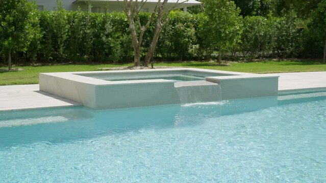 Modern outdoors swimming pool with clear blue water. Concrete structure in middle possibly a fountain or island feature and lush greenery in background