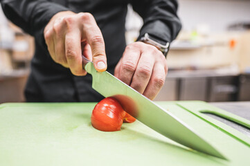 A person slices a tomato on a green cutting board using a kitchen knife.